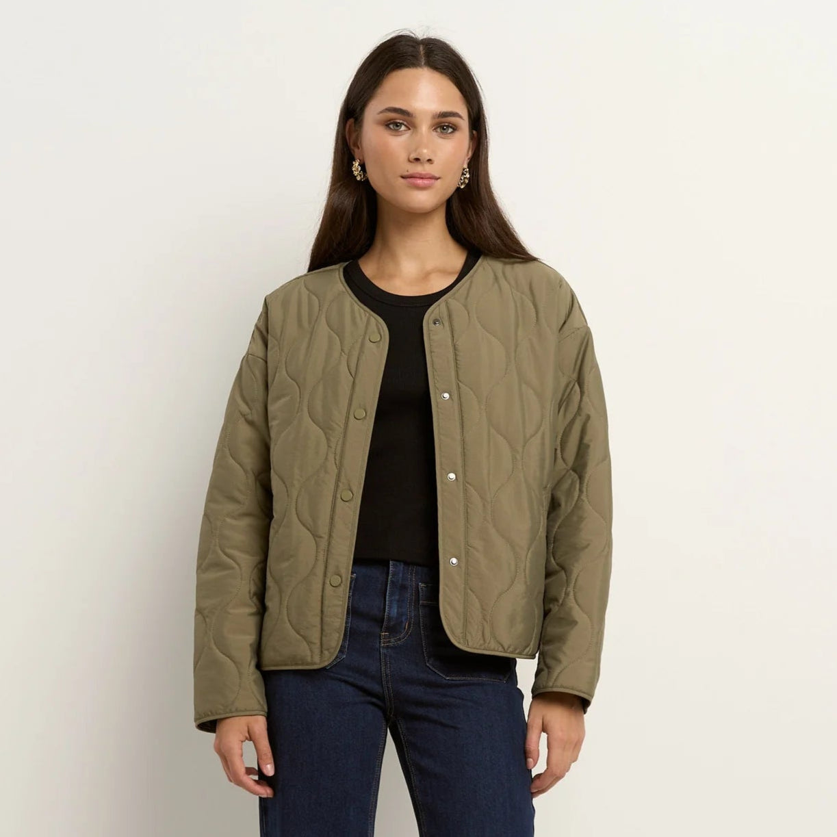 Woman wearing a green quilted jacket, black top, and blue jeans on a white background